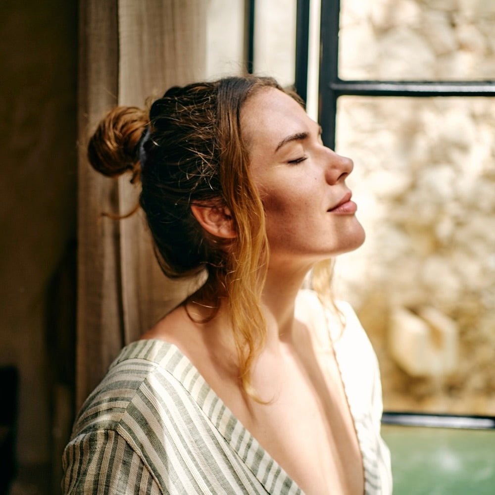 Woman with a bun in her hair, wearing a striped off-shoulder top, standing by a window.